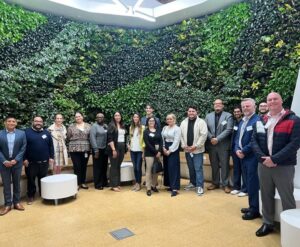 A line of individuals in an office setting, in front of a wall of live plants. 