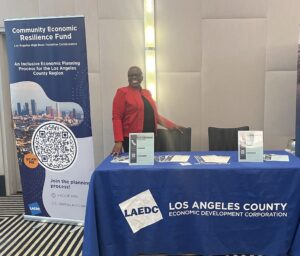 A woman is smiling while standing next to a table with informational items at the LAEDC 88 Cities Summit 2023